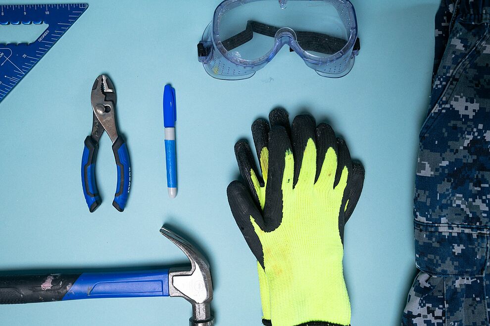Arbeitssicherheit: Persönliche Schutzausrüstung mit Schutzhandschuhen, Schutzbrille und Werkzeugen auf Arbeitsfläche.