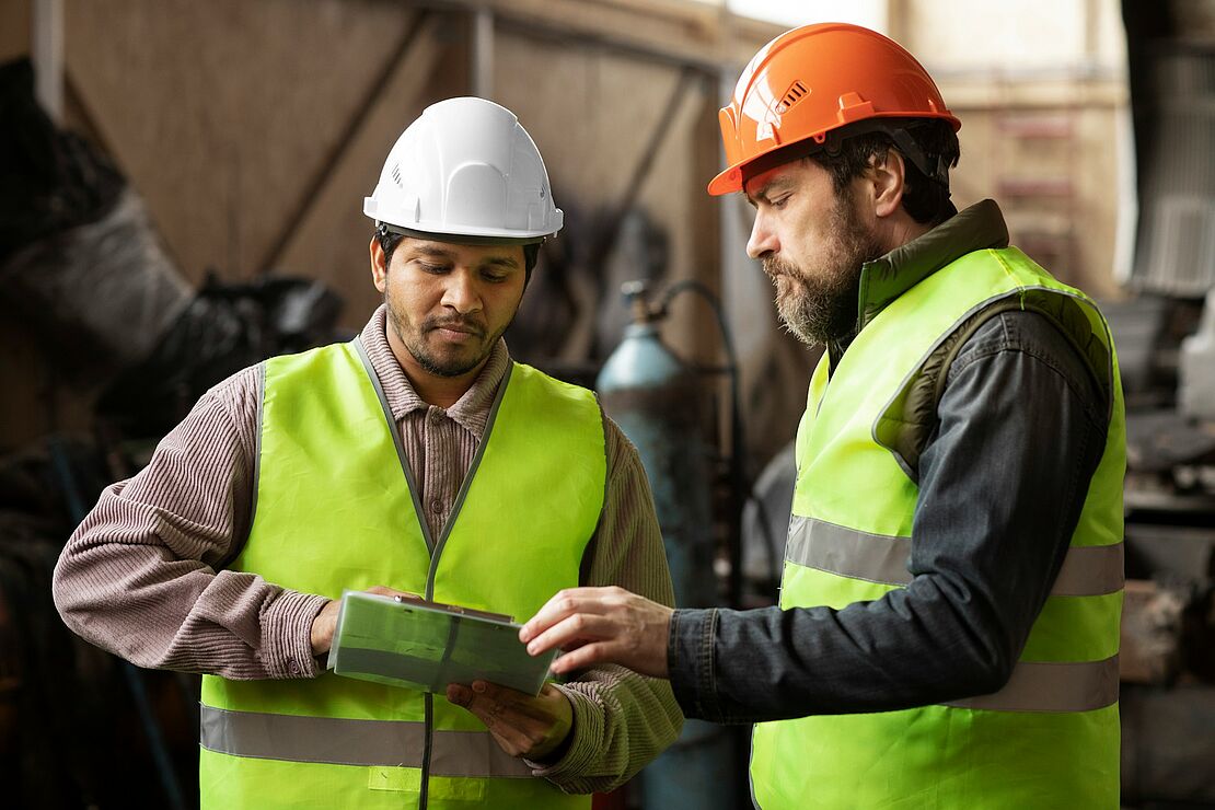 Arbeitssicherheit in der Industrie: Zwei Mitarbeitende mit Schutzhelm und Warnweste prüfen Arbeitsunterlagen in einer Produktionshalle.