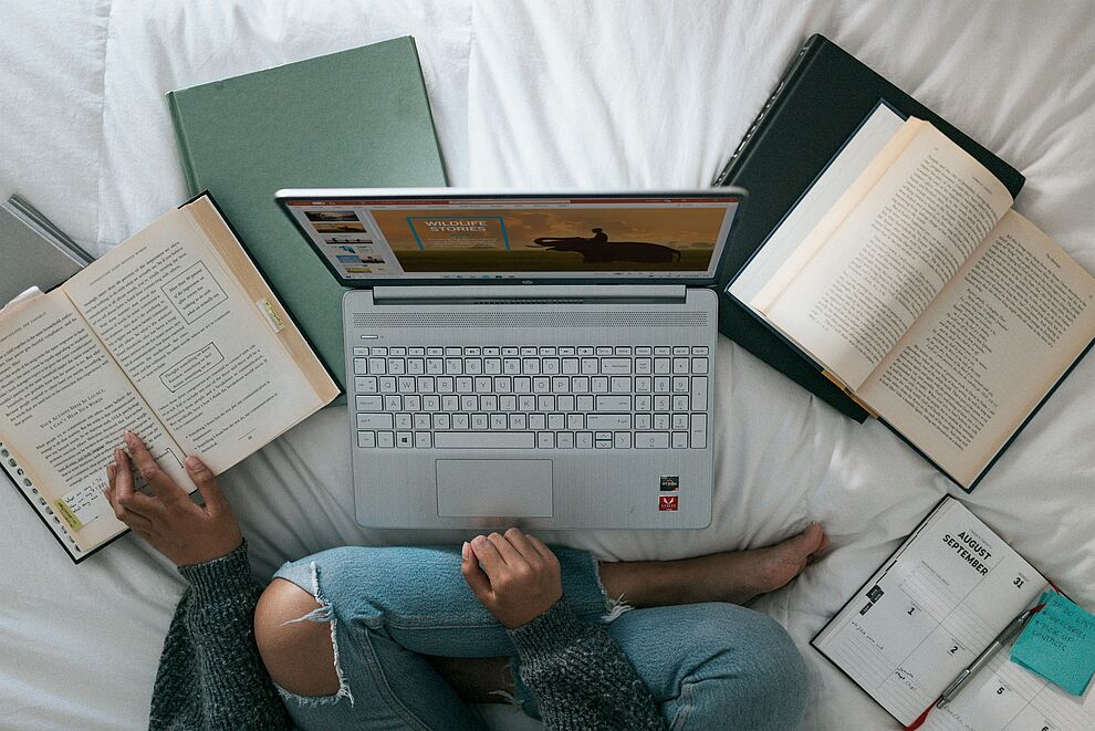 Laptop liegt zwischen mehreren offenen Büchern und Notizen auf einem Bett, dargestellt ist eine Lernsituation für eine Online Weiterbildung