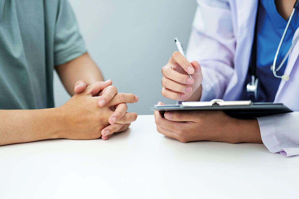 Zwei am Tisch sitzende Personen, von der eine Person einen Arztkittel trägt und etwas auf einem Klemmbrett notiert