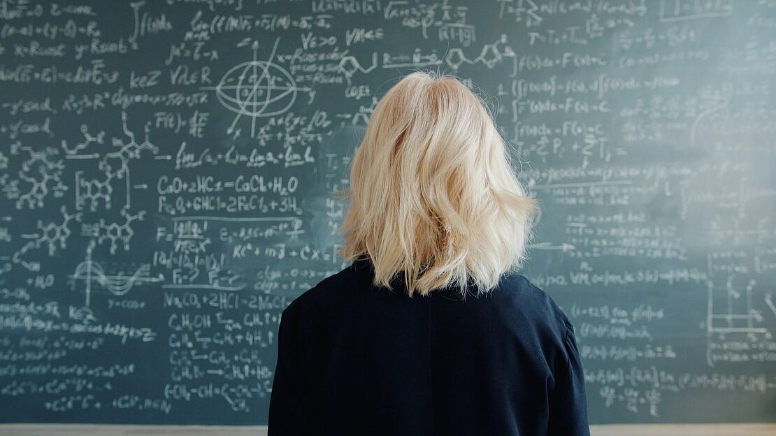 Person von hinten vor einer Tafel mit mathematischen und chemischen Formeln als Symbol für komplexes Fachwissen und die Bedeutung verständlicher Grundlagen.