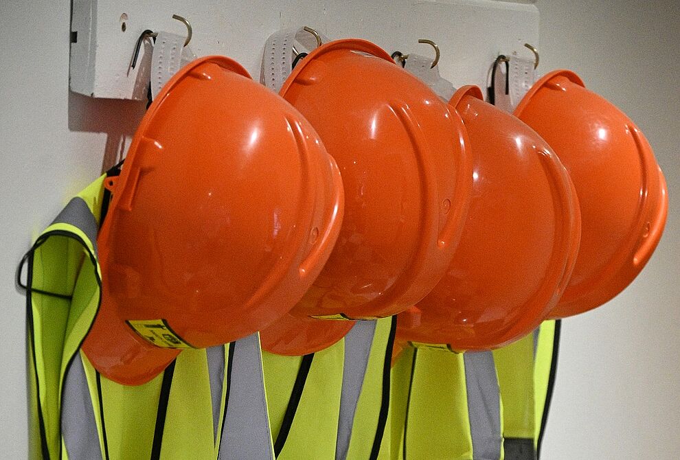 Reihe von orangenen Schutzhelmen und reflektierenden Warnwesten, wie sie im Arbeitsumfeld von Fachkräften für Arbeitssicherheit verwendet werden.