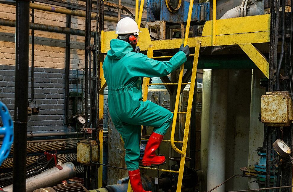 Eine Person mit Schutzbekleidung steigt in einer Anlage mit Rohrleitungen und Maschinen eine Leiter hinauf. Die Szene zeigt typische Arbeitsumgebungen, in denen Maßnahmen zum Gewässerschutz durchgeführt und überwacht werden.