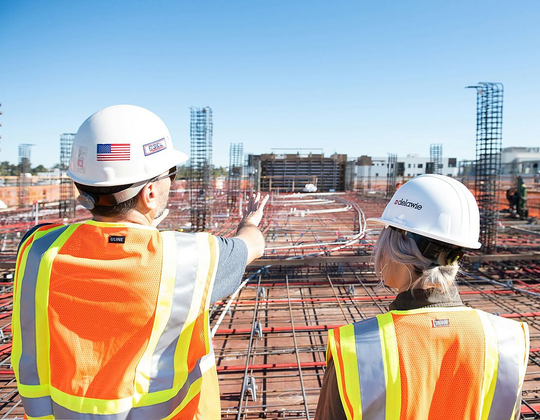 ALT- Text: Zwei Personen mit Schutzhelmen und Warnwesten betrachten eine große Baustelle – eine typische Situation für die Arbeit einer Fachkraft für Arbeitssicherheit.