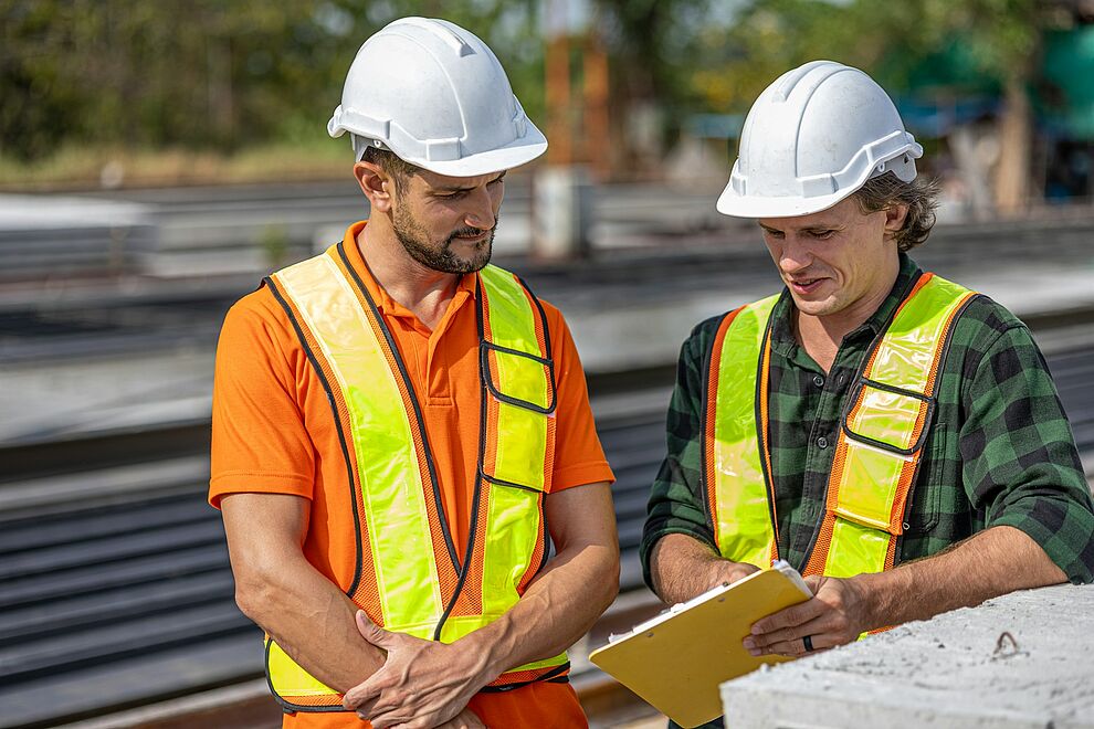 Zwei Beschäftigte mit Schutzhelm und Warnwesten prüfen Unterlagen zu Arbeitssicherheit auf einer Baustelle