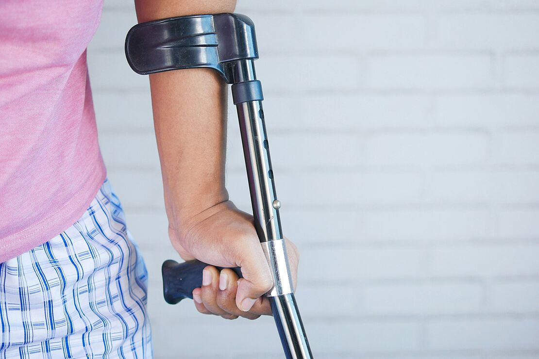 Eine Person stützt sich mit einer Unterarmgehstütze ab und steht vor einer weißen Wand als Beispiel für Verletzungsfolgen und Bedeutung präventiver Maßnahmen