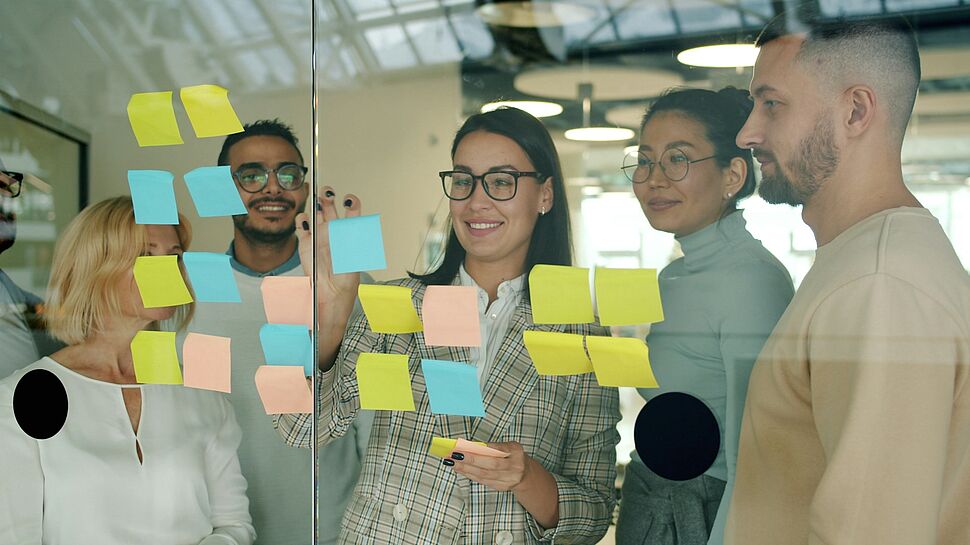 ALT-Text: Mehrere Personen stehen vor einer Glaswand mit farbigen Haftnotizen und besprechen gemeinsam Ideen und Maßnahmen. Das Bild steht im Kontext eines Workshops zum HSE-Management.