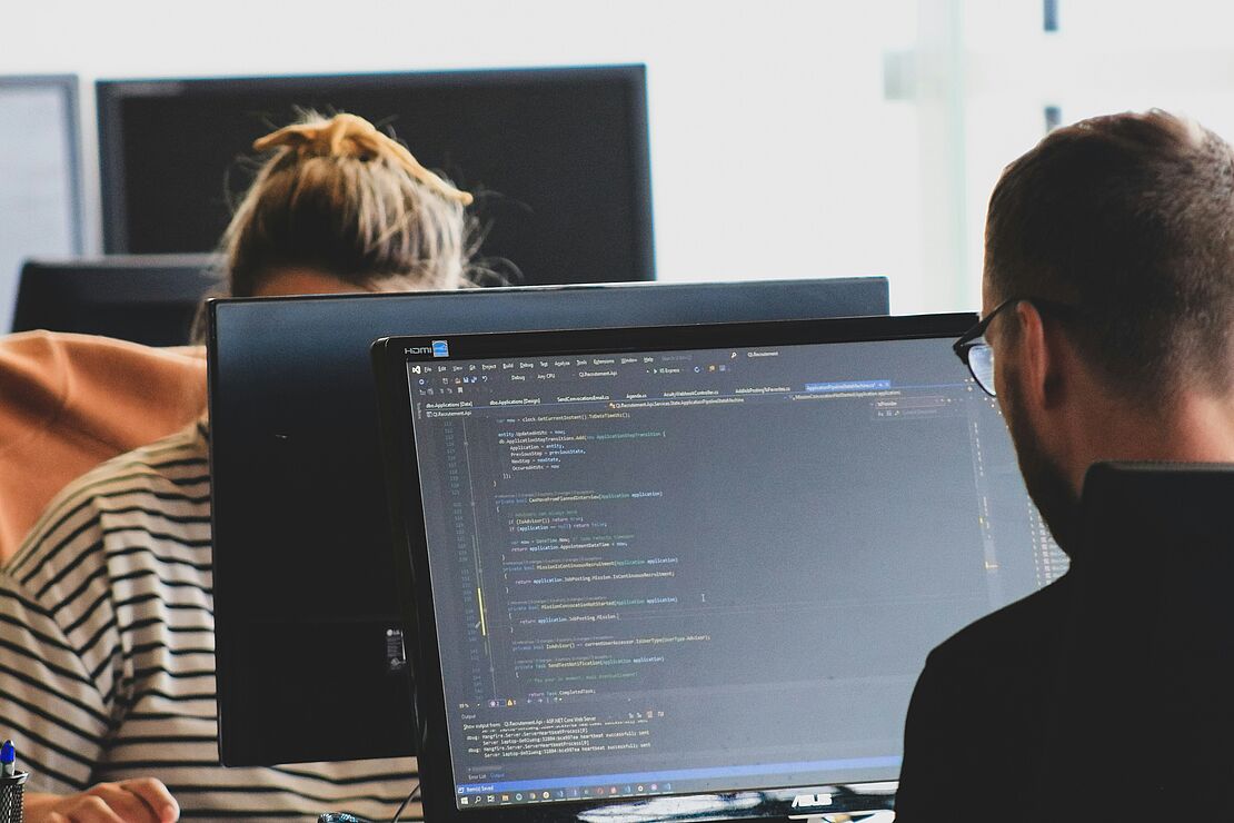 Personen arbeiten an einem Schreibtisch mit Computerbildschirmen und Dokumenten zur Steuerung und Integration von Managementsystemen im Unternehmen