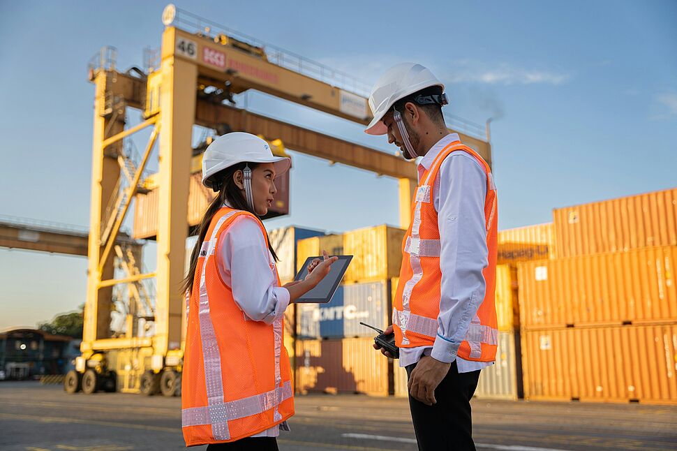 Zwei Beschäftigte mit Schutzhelm und Warnwesten besprechen Arbeitsschutzmaßnahmen auf einem Containerterminal